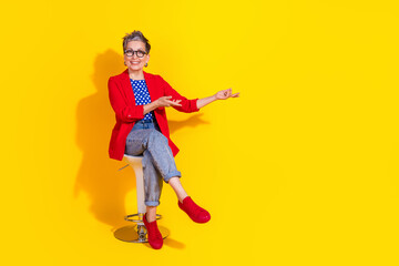 Stylish mature woman in a red jacket sitting on a stool in front of a bright yellow background,...