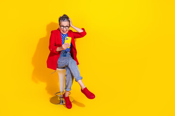 Stylish middle-aged woman in red blazer sitting on stool with smartphone against vibrant yellow background