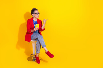 Stylish mature woman in bright casual fashion sitting against yellow background, holding smartphone with a smile