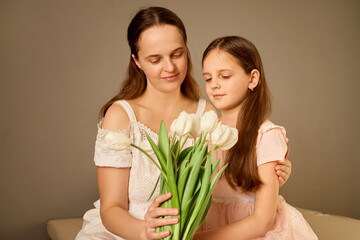 A woman and a young girl sit closely together, gazing at a bouquet of white tulips. The soft...