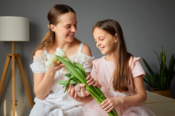Obraz premium A mother and her daughter smile as they hold white tulips together in a cozy indoor setting. Natural light fills the room, creating a warm and inviting atmosphere
