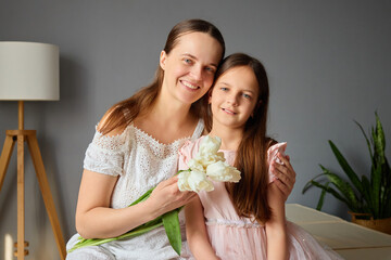 Obraz premium A mother and her daughter smile brightly as they sit together on a comfortable sofa, enjoying a moment with fresh flowers in a warm, inviting room filled with natural light