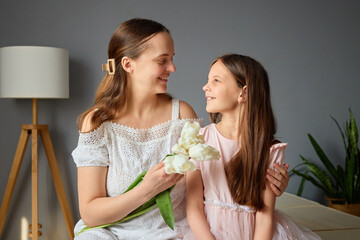 A mother smiles at her daughter while holding a bouquet of white flowers. They enjoy a warm moment together in a cozy living room filled with greenery and soft light