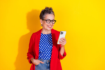 Cheerful mature woman using smartphone in vibrant outfit against yellow background, reflecting happiness, style, and technology.