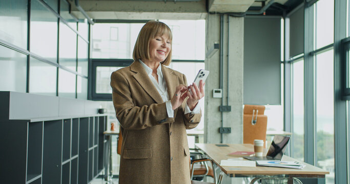 A mature businesswoman using a smartphone in a modern office setting, checking messages or emails while standing confidently, showcasing professionalism and digital connectivity