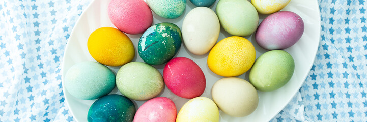 Multi-colored Easter eggs in an oval dish on a blue tablecloth, top view, Happy Easter holiday banner