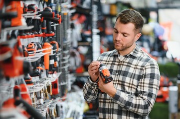 Portrait of young bearded man buying modern electric drill while standing in tool. Hardware...