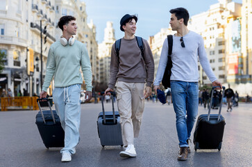 Happy male tourists are walking down a city street, pulling rolling suitcases and talking