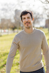 A middle-aged confident handsome man with light stubble smiles slightly in a sunlit park. Looking at camera. Blurred urban background with green grass. Vertical, soft focus, natural light