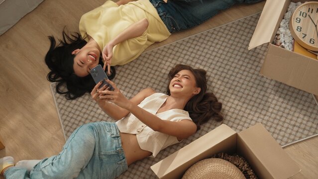 Lesbian lover looking at phone while laughing together and lying down at living room floor with cardboard box scattering. Happy couple checking at smart phone while spending time together. Myrmidon.