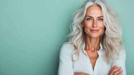 A beautiful older woman with flowing gray hair smiles confidently while posing in front of a serene green background, exuding elegance and charm.