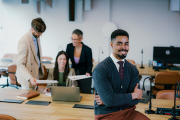 Obraz premium Portrait of confident smiling black businessman sitting leaning on desk in office, posing with folded arms and looking at camera, colleagues working in blurred background