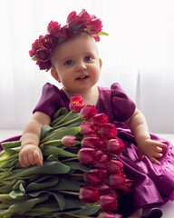 A portrait of a cheerful, beautiful one-year-old girl stands in a burgundy dress at the window of the house. He wears a wreath of flowers on his head