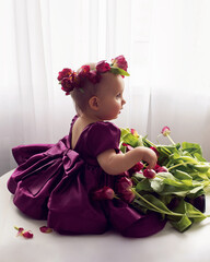 A portrait of a cheerful, beautiful one-year-old girl sit in a burgundy dress at the window of the house. He wears a wreath of flowers on his head