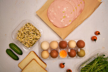 On the table are farm eggs of different colors, pieces of cooked sausage, microgreens, toasted white bread and vegetables, top view on a light-colored background