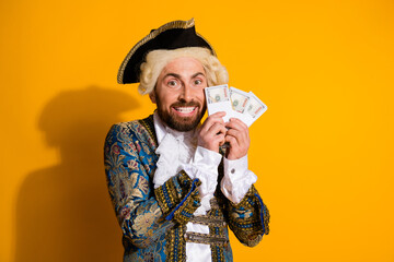 Elegant man in baroque costume holding money with a cheerful expression against yellow background