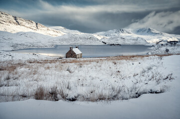 Typical Scottish stone house on the shore of Loch stack lake during winter day in a snow covered landscape, yellow grass in foreground and snow capped mountains in background, Assynt, Scotland © Ji