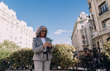 Sophisticated mature woman in fashionable suit looks at mobile phone with smile, checking messages...