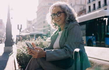 Confident senior businesswoman sits on bench in the city, smiling while holding her smartphone....