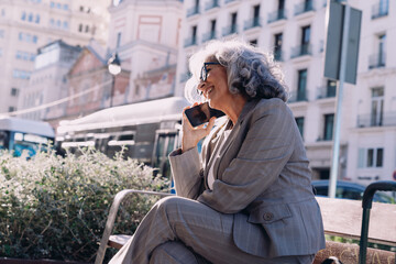 Fototapeta premium Happy senior businesswoman in suit enjoys mobile phone conversation while sitting on bench in city. Female looks relaxed and confident, reflecting digital connectivity, leadership, work-life balance