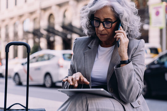 Senior businesswoman in gray suit sits outdoors, engaged in a phone call while working on her tablet. Female focused expression suggests multitasking and efficiency in a busy urban setting