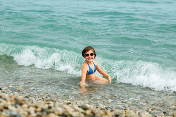 A small, beautiful girl in a blue swimsuit and sunglasses, swimming on the beach