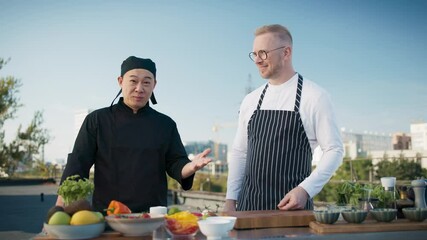 Multiethnic male chefs vloggers smiling while preparing food on rooftop. Content creators filming outdoor cooking session with fresh ingredients and city skyline in background. Vlog of restaurant.