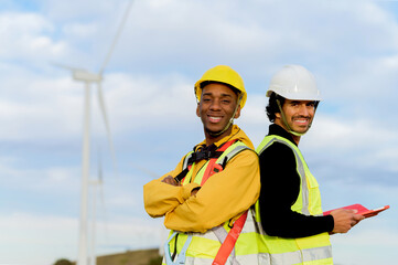 Two multi ethnic engineers are smiling in a wind turbine farm, wearing safety vests and helmets,...