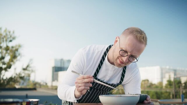 Caucasian male chef adding finishing touches to dish, using precision to enhance the presentation. The backdrop of cityscape and rooftop setting, combining culinary artistry with urban charm.