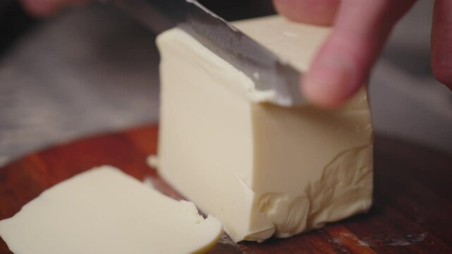 Butter Cutting Process Closeup. Chef Slicing Fresh Butter Block on Wooden Board - Food Preparation Macro Shot. Kitchen Cooking Ingredient Being Cut into Thin Slices. Culinary Dairy Product Preparation