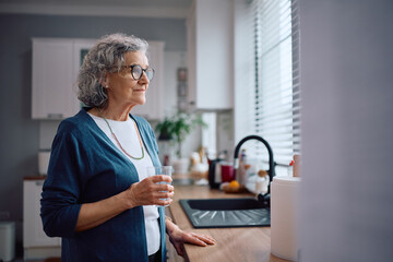 Thoughtful elderly woman having a glass of water in the kitchen.