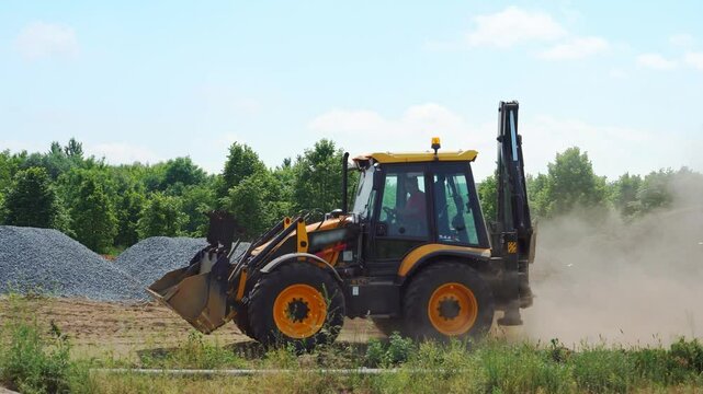 An excavator with a bucket drives along a dusty road against the background of an industrial plant for the extraction and processing of crushed stone.