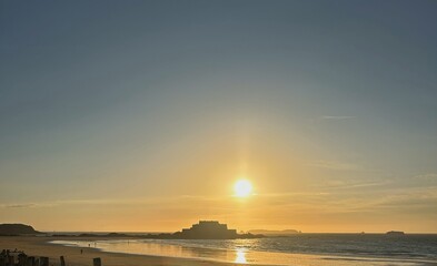 Golden hour at Saint-Malo, the sun setting behind the historic fort, casting a warm glow over the beach. The wooden breakwaters stand tall, weathered by time and tides, framing the tranquil seascape.