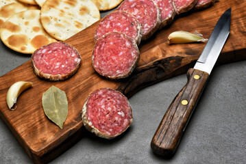 Slices of a salami roll on a cutting board with crackers, spices and herbs, and a knife.