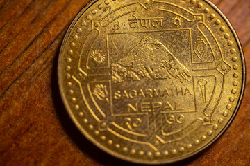 A close-up of a Nepalese coin depicting Mount Sagarmatha (Everest) with intricate engravings. The gold-toned coin rests on a wooden surface, highlighting its fine details and texture.