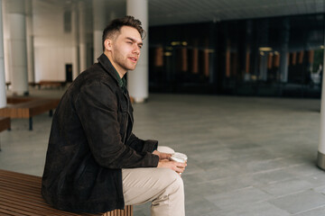 Side view of pensive young man sitting on wooden bench in contemporary city environment, holding...
