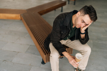 High-angle view of frustrated pensive man sitting on street bench, holding coffee cup and resting...