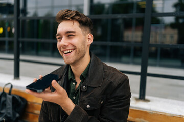 Close-up of cheerful young male recording voice message on smartphone while sitting on bench in city street, enjoying moment of relaxation during workday. Concept of modern successful city lifestyle.