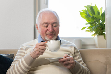 Happy senior man holding cup of coffee while relaxing sofa home. Smiling mature older man drinking tea looking at camera while sitting on cozy sofa relaxing in modern kitchen living room
