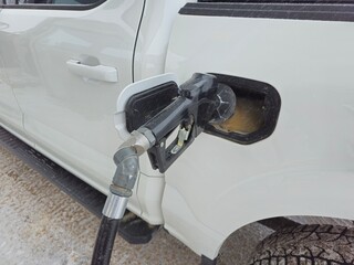 A fuel pump nozzle inserted into the fuel tank of a white pickup truck. The handle is brass and is...