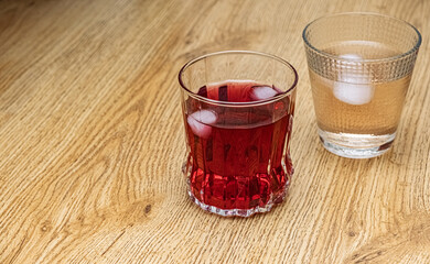 Refreshing drinks with Ice. Two glasses, one filled with a red beverage and ice cubes, and the other with a clear liquid and ice, are positioned on a light wooden table.
