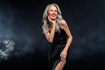 Mature woman in a glamorous black dress with silver hair smiling confidently in a dark setting