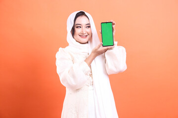 Smiling Muslim woman in hijab presents a smartphone with green screen display, happy to communicate...