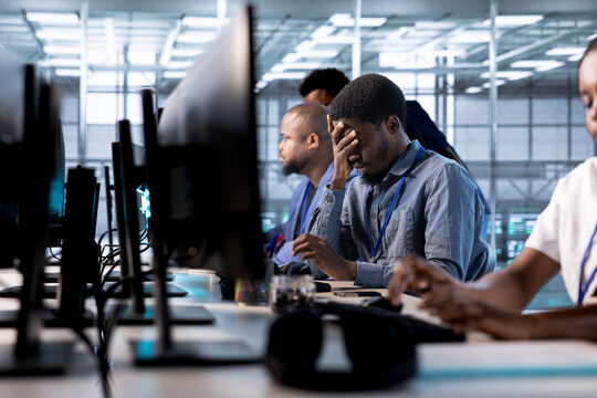 Tired engineer feeling headache while reconfiguring server farm to optimize it for data processing requirements. Stressed man working overtime to ensure data center gear operates efficiently - Powered by Adobe