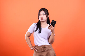 Woman with a smartphone looks annoyed while standing against an orange background
