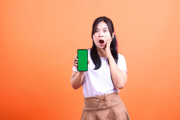 Naklejka premium A woman asia wearing a white shirt and brown skirt holding a smart phone device with a green screen of a surprised expression with an open mouth and one hand raised isolated on an orange background
