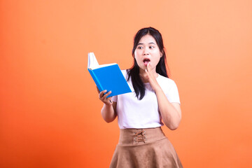 Fototapeta premium A woman reading a blue book with a surprised expression, wearing a white shirt and brown skirt isolated on orange background
