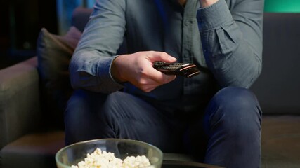 Close up of man on couch using remote control to browse though movies available on streaming service. Focus on remote control used by homeowner in apartment switching channels on TV, camera A