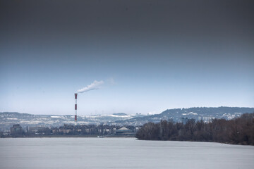 Fototapeta premium Industrial chimney emitting smoke causing visible air pollution over the frozen Danube river during wintertime in Belgrade, Serbia, illustrating environmental issues and industrial impact.