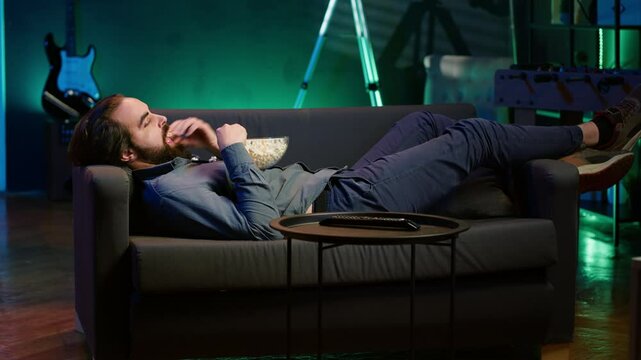 Man procrastinating at home, laying down on couch, munching on snacks, watching TV. Bored person living sedentary lifestyle, lounging on couch, wasting time watching TV, eating popcorn, camera A